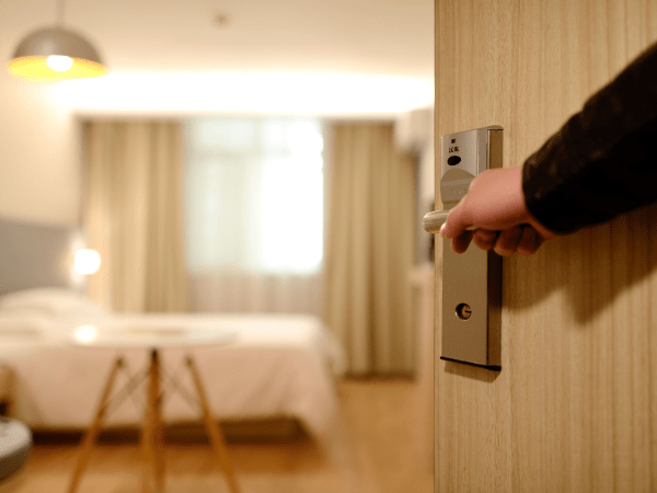 A hand opens a wooden door to a well-lit hotel room with a bed, curtains, and a modern light fixture.